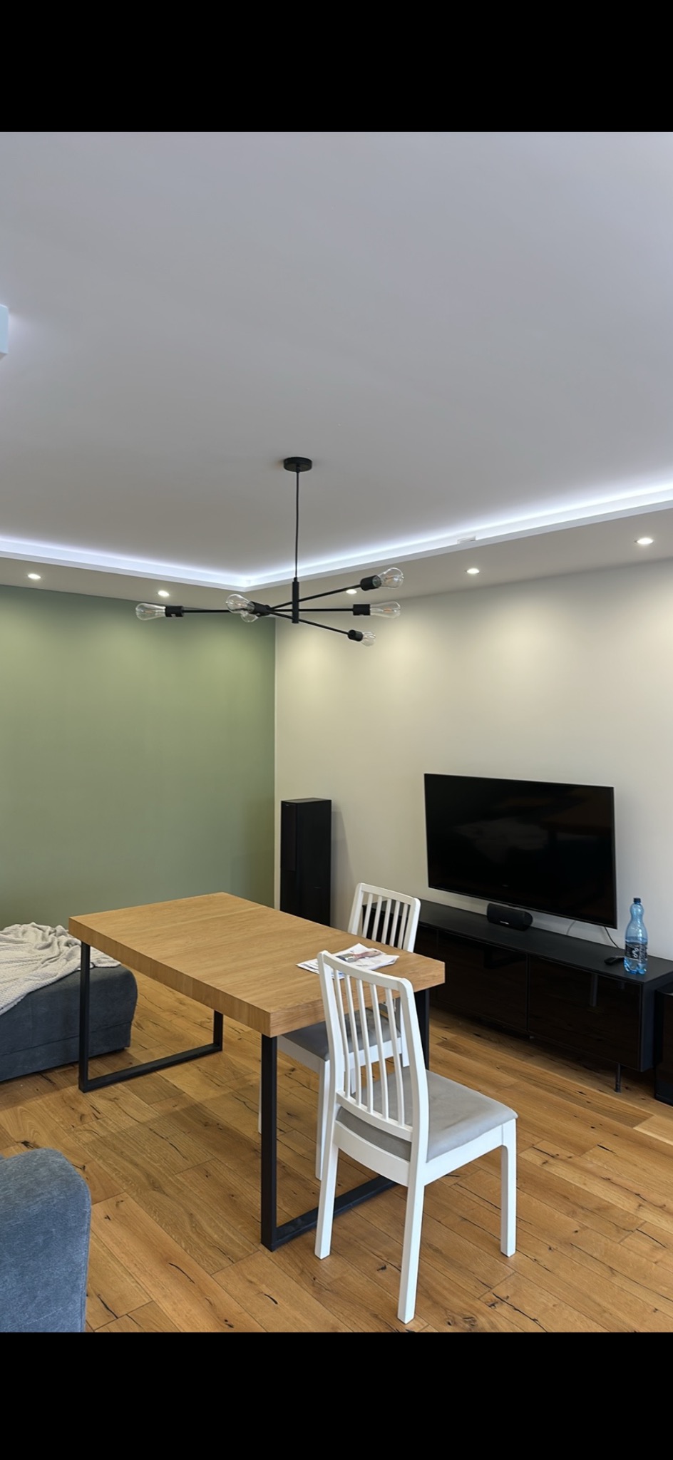 Jasne wnętrze: drewniana podłoga, stół z czarnymi nogami, białe krzesła, minimalistyczny żyrandol. Ściana w odcieniach zieleni i beżu. Nowoczesny design.