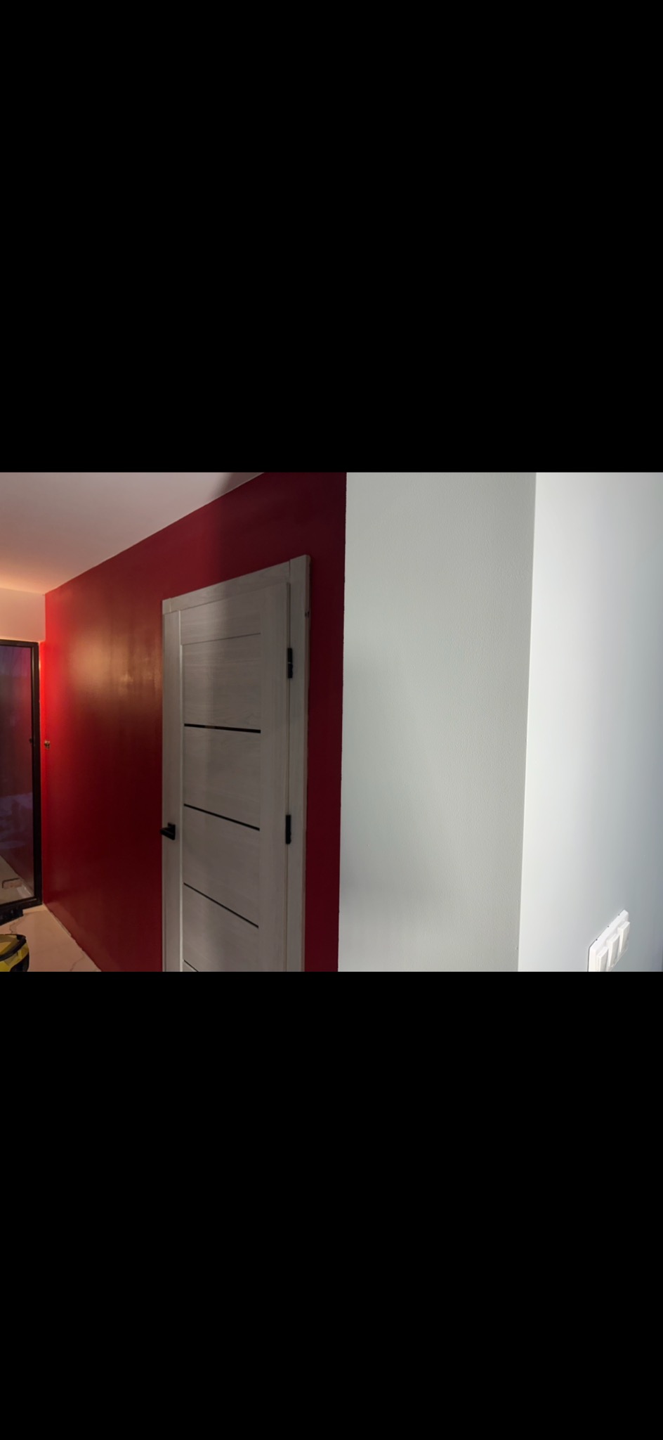 Ściana pomalowana na czerwono z zamontowanymi drzwiami z poziomymi paskami, sąsiadująca z białą ścianą. Widoczny fragment podłogi i włącznik światła.