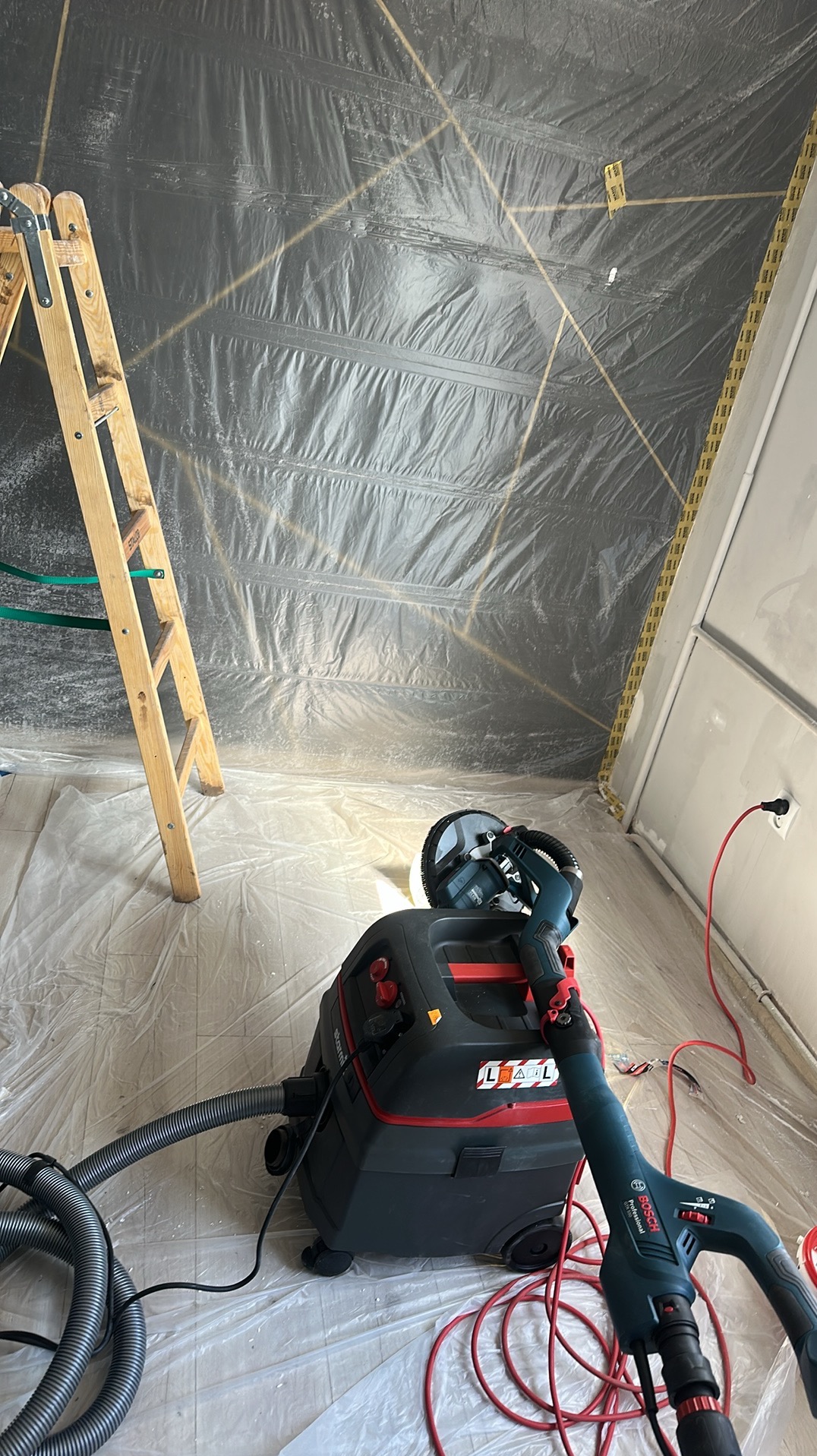 Przygotowane pomieszczenie do prac wykończeniowych: folia ochronna, drabina i szlifierka do gładzi z odkurzaczem przemysłowym. Widok z góry na narzędzia.