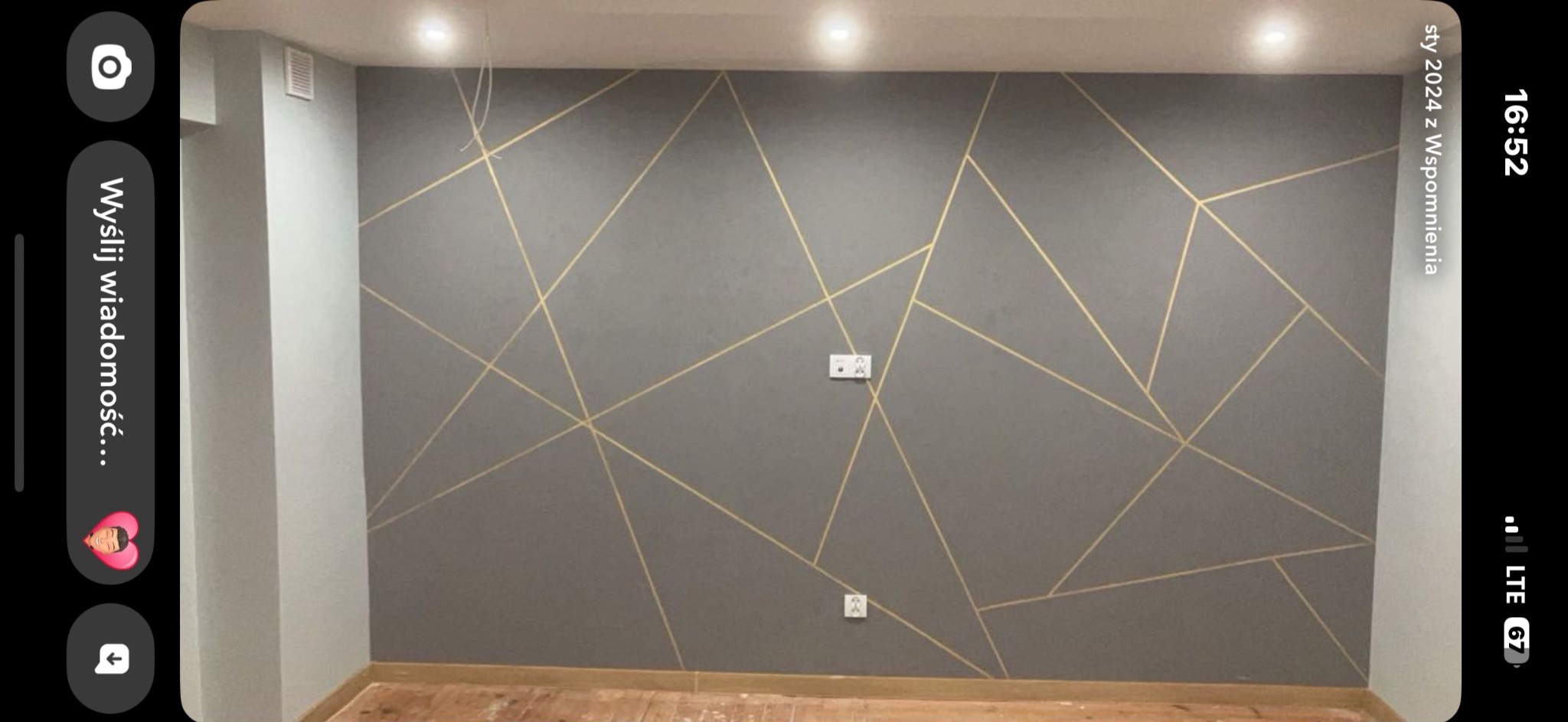 Szara ściana z geometrycznym wzorem z złotych linii, widoczne gniazdka elektryczne. Podłoga drewniana, jasne oświetlenie sufitowe.