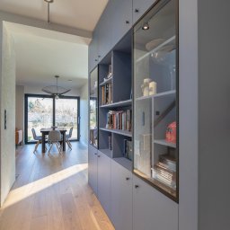 ZPHU Budziński&Motyka s.c. - Niebieska zabudowa meblowa z przeszklonymi witrynami w korytarzu, widok na jadalnię z drewnianą podłogą i dużym oknem. Nowoczesny design, stonowana kolorystyka.