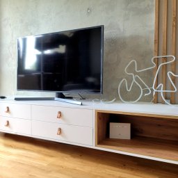 ZPHU Budziński&Motyka s.c. - Biała szafka RTV z szufladami i skórzanymi uchwytami, na drewnianej podłodze, z dekoracją w kształcie roweru. Minimalistyczny design, betonowa ściana w tle.