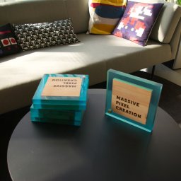 ZPHU Budziński&Motyka s.c. - Dwa unikalne, akrylowe bloki z drewnianymi wstawkami z napisem 'Massive Pixel Creation' na okrągłym stoliku kawowym, w tle sofa z poduszkami o tematyce retro gier.