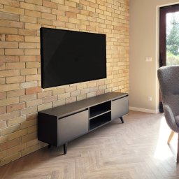 ZPHU Budziński&Motyka s.c. - Szara szafka RTV z czarnymi nóżkami pod telewizorem na ceglanej ścianie. Parkiet ułożony w jodełkę. Minimalistyczny design, nowoczesne wnętrze.