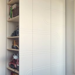 ZPHU Budziński&Motyka s.c. - Biała szafa z półkami, wypełniona zabawkami, z dekoracyjnym frezem na frontach, stojąca na drewnianych nóżkach w pokoju dziecięcym.