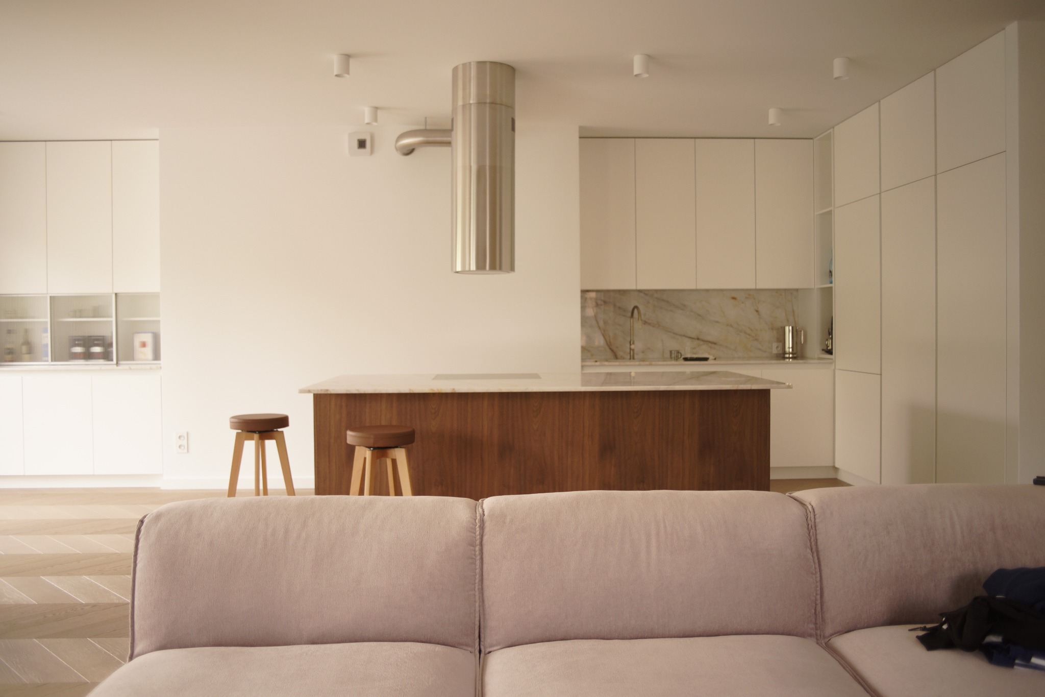 Nowoczesna kuchnia z wyspą i marmurowym blatem, widziana zza sofy. Minimalistyczne białe szafki i drewniane wykończenia tworzą elegancką przestrzeń.
