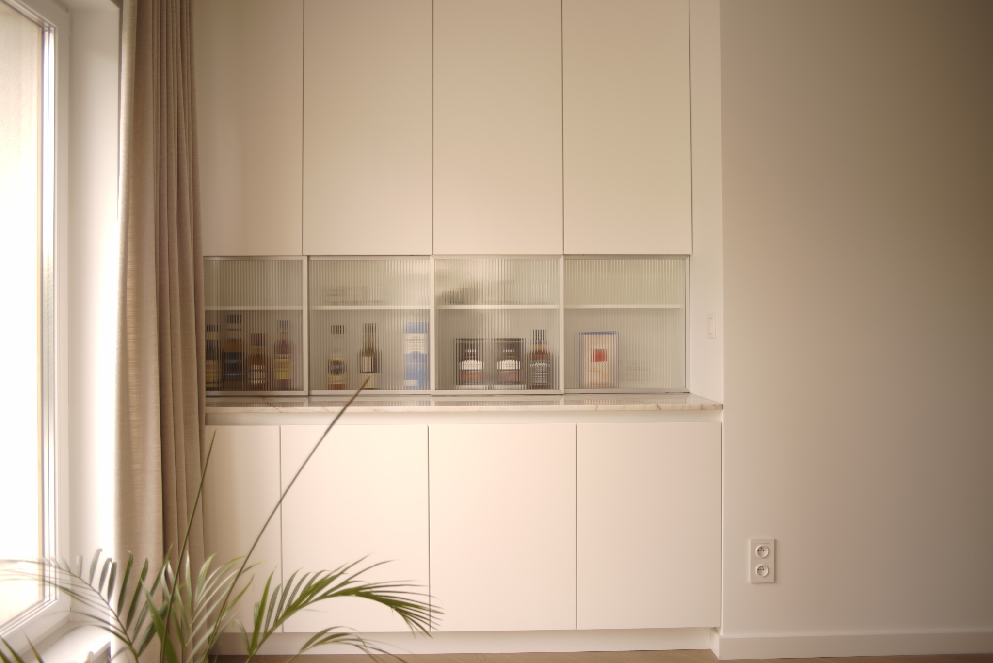 Biała zabudowa kuchenna z ryflowanymi szybami, eksponująca butelki i dekoracje. Minimalistyczny design, neutralna kolorystyka, roślina doniczkowa w lewym dolnym rogu.