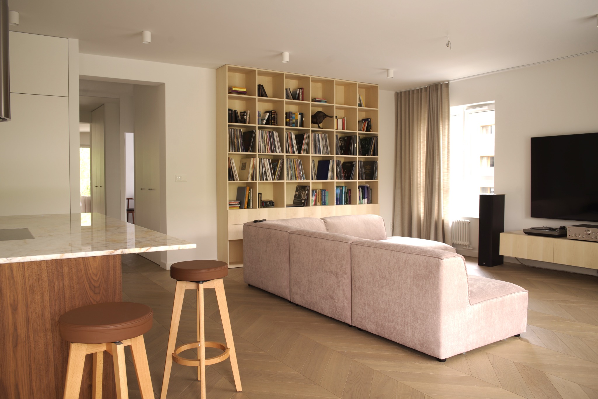 Jasna, nowoczesna przestrzeń: beżowa sofa, regał z książkami, drewniane krzesła barowe przy wyspie kuchennej z marmurowym blatem i parkiet w jodełkę.