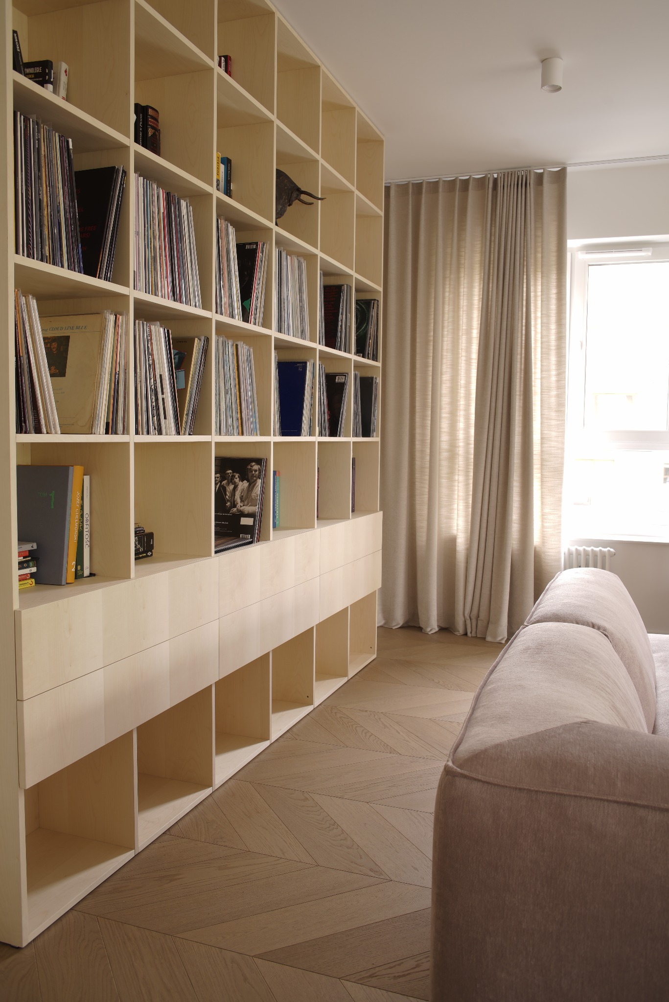 Jasna, drewniana szafa z półkami i szufladami, wypełniona książkami i płytami, ustawiona w salonie z parkietem w jodełkę i beżową sofą. Minimalistyczny design.