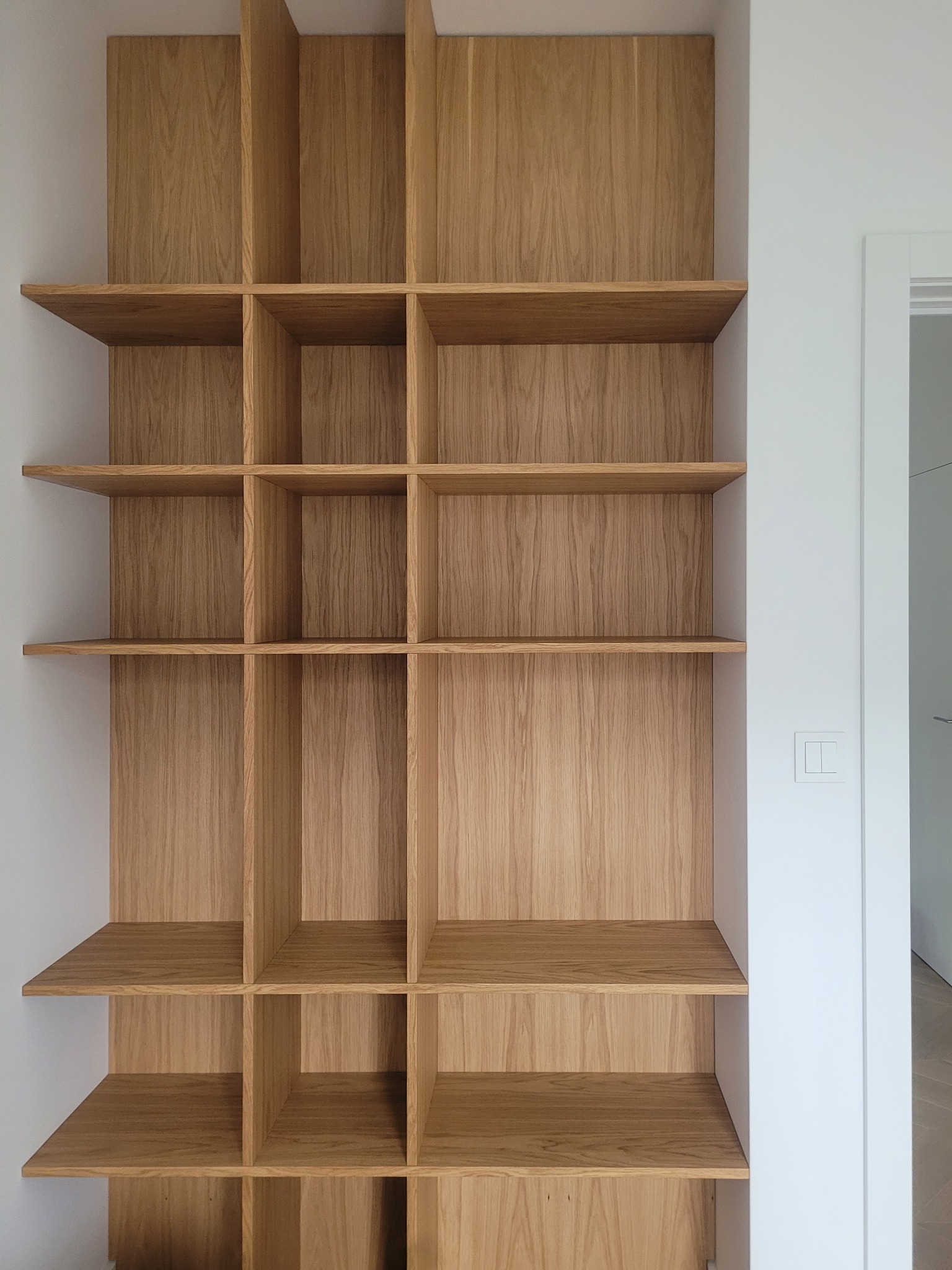 Półki z jasnego drewna wnękowe, minimalistyczny design. Widoczne detale wykończenia i naturalna tekstura drewna. Białe ściany i fragment drzwi w tle.