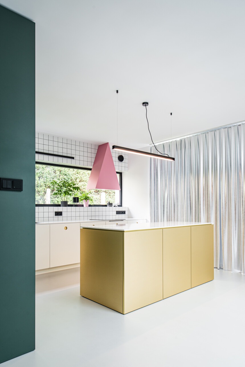 Nowoczesna kuchnia z wyspą w kolorze złota, różowym okapem i białymi szafkami. Minimalistyczny design, okno z widokiem na zieleń, srebrne zasłony. Nietypowe oświetlenie.