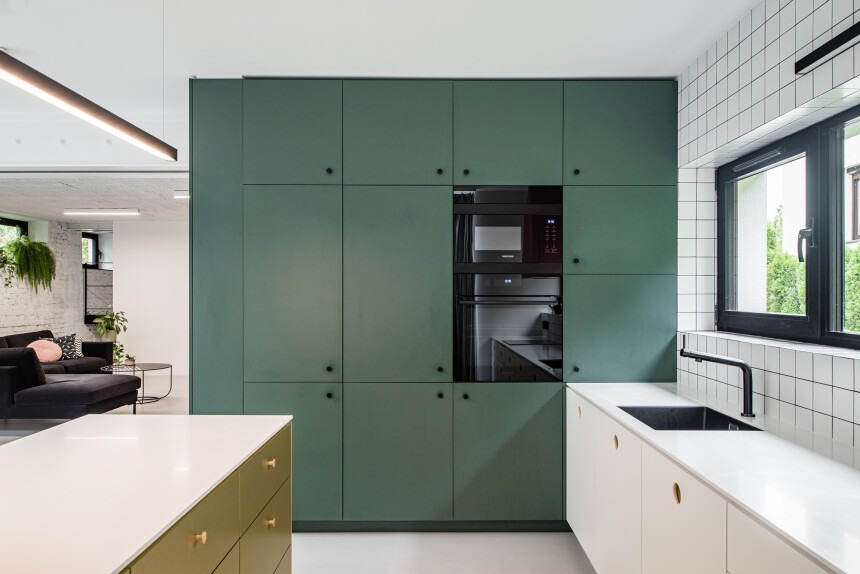 Nowoczesna kuchnia z zielonymi szafkami, czarnym piekarnikiem i mikrofalówką. Biały blat i płytki. W tle salon z czarną sofą i roślinami.