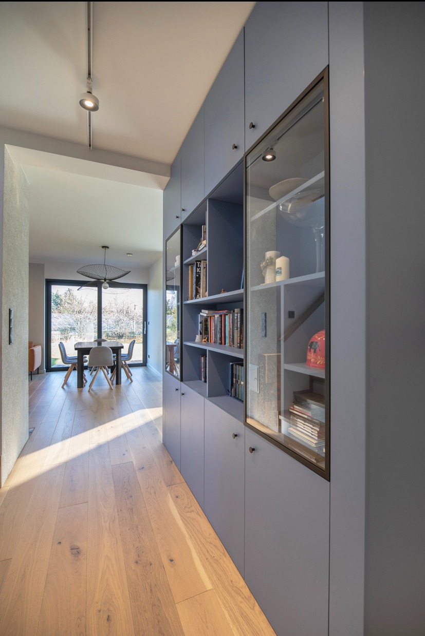 Niebieska zabudowa meblowa z przeszklonymi witrynami w korytarzu, widok na jadalnię z drewnianą podłogą i dużym oknem. Nowoczesny design, stonowana kolorystyka.