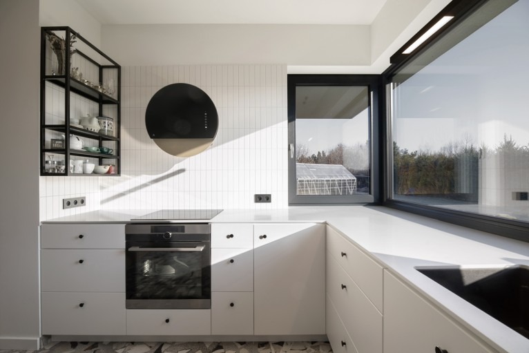 Nowoczesna, jasna kuchnia z białymi szafkami, czarnymi uchwytami i okrągłym okapem. Duże okno z widokiem na szklarnię. Metalowy regał z naczyniami.
