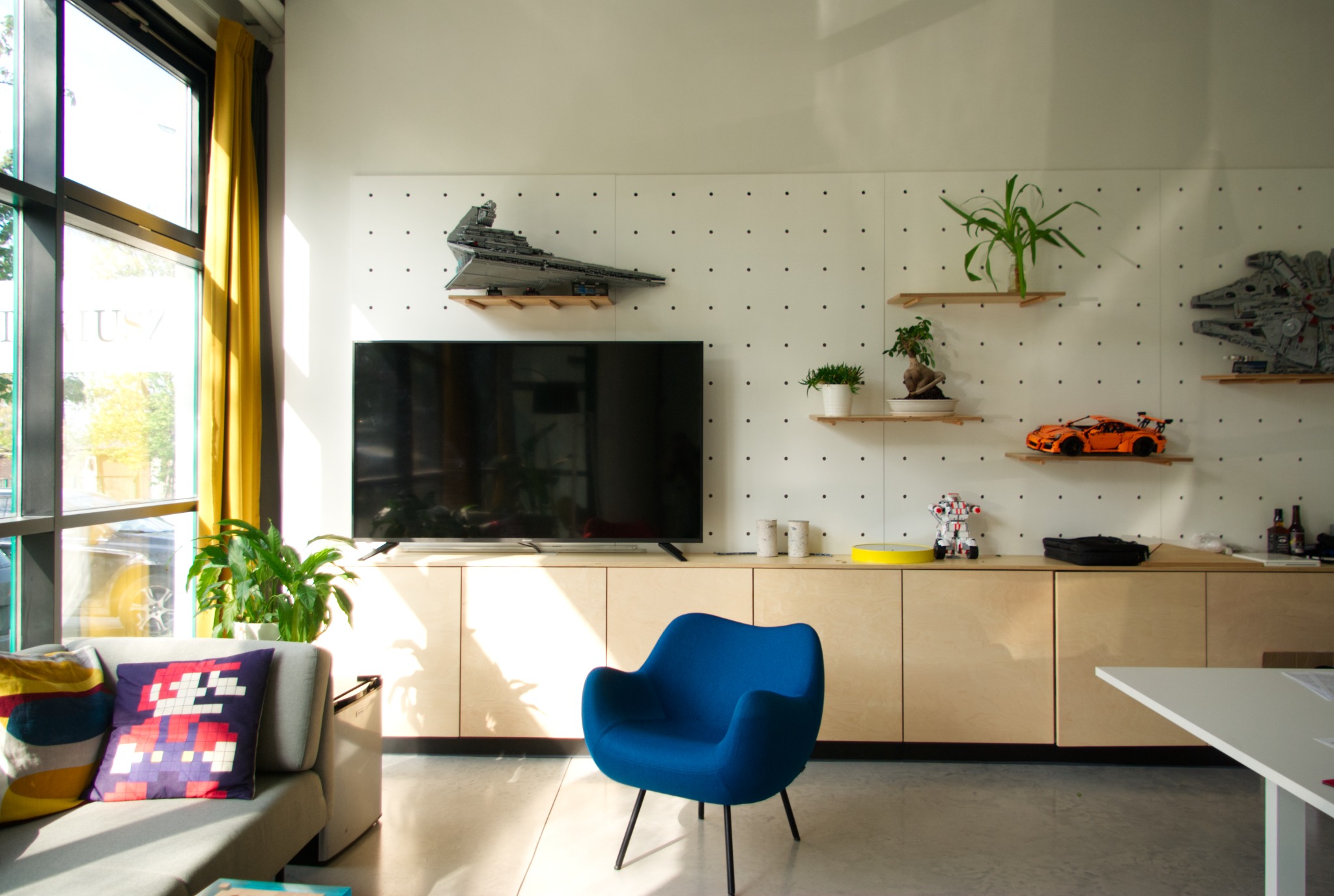 Jasne wnętrze pokoju z szafką RTV z jasnego drewna, na której stoi telewizor. Na ścianie półki z dekoracjami. W pokoju sofa i niebieski fotel.