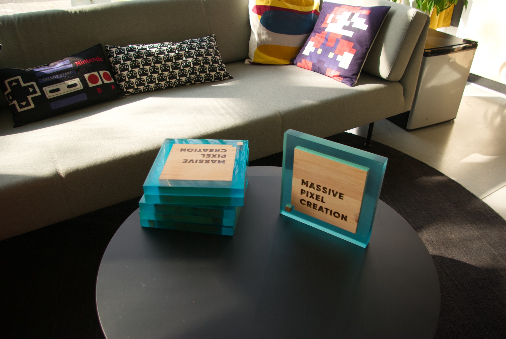 Dwa unikalne, akrylowe bloki z drewnianymi wstawkami z napisem 'Massive Pixel Creation' na okrągłym stoliku kawowym, w tle sofa z poduszkami o tematyce retro gier.