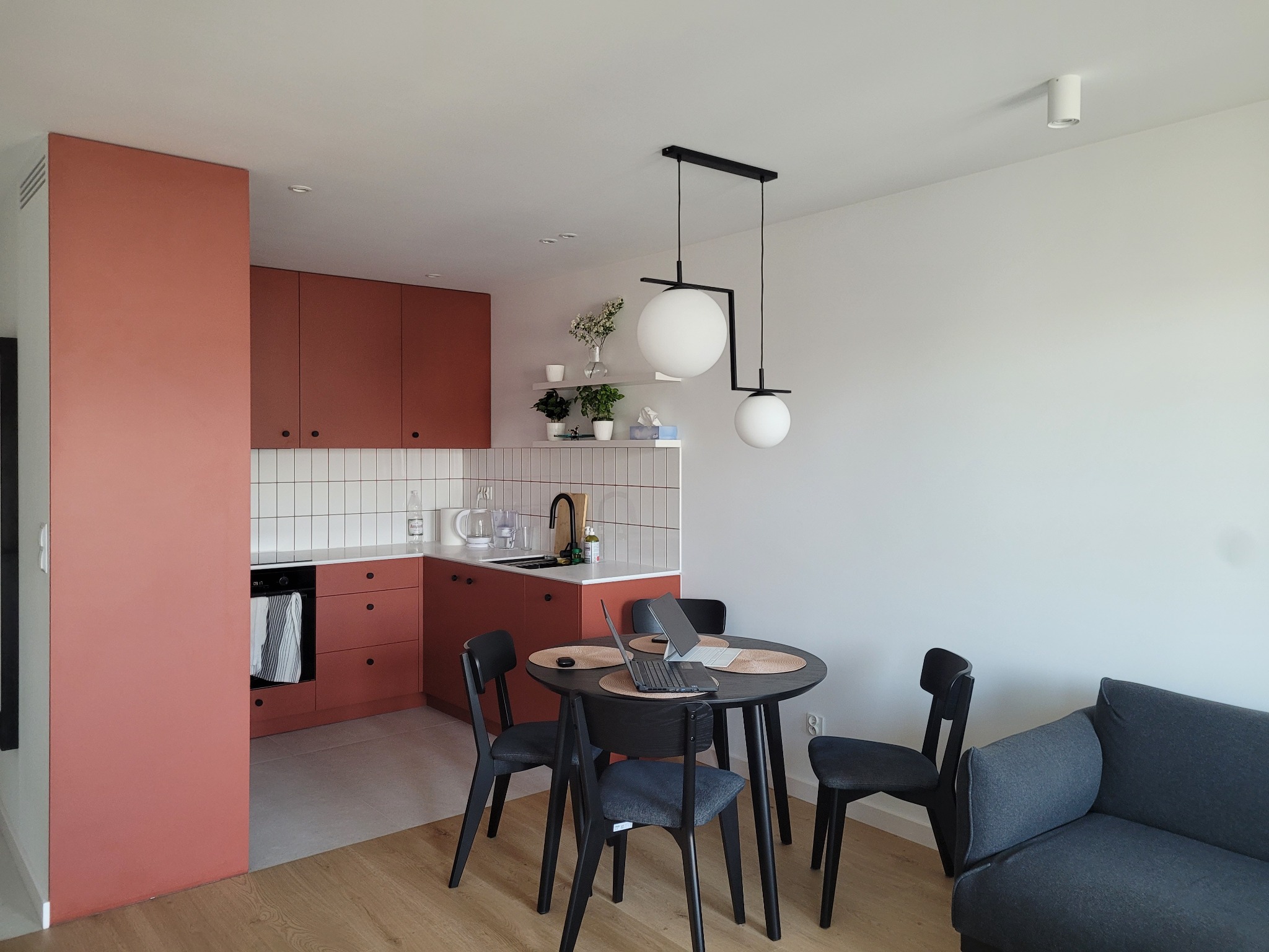 Nowoczesna kuchnia w odcieniach terakoty z białymi płytkami, okrągły stół z laptopem i czarnymi krzesłami, minimalistyczna lampa wisząca i szara sofa w tle.