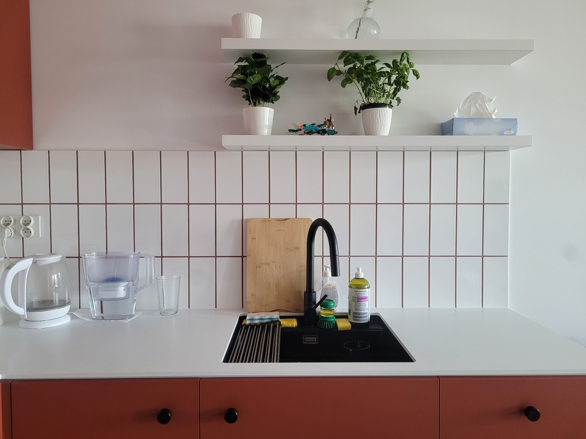 Nowoczesna kuchnia z terakotowymi szafkami, białym blatem i czarnym zlewem. Na ścianie białe płytki i półki z roślinami. Minimalistyczny design, jasne wnętrze.