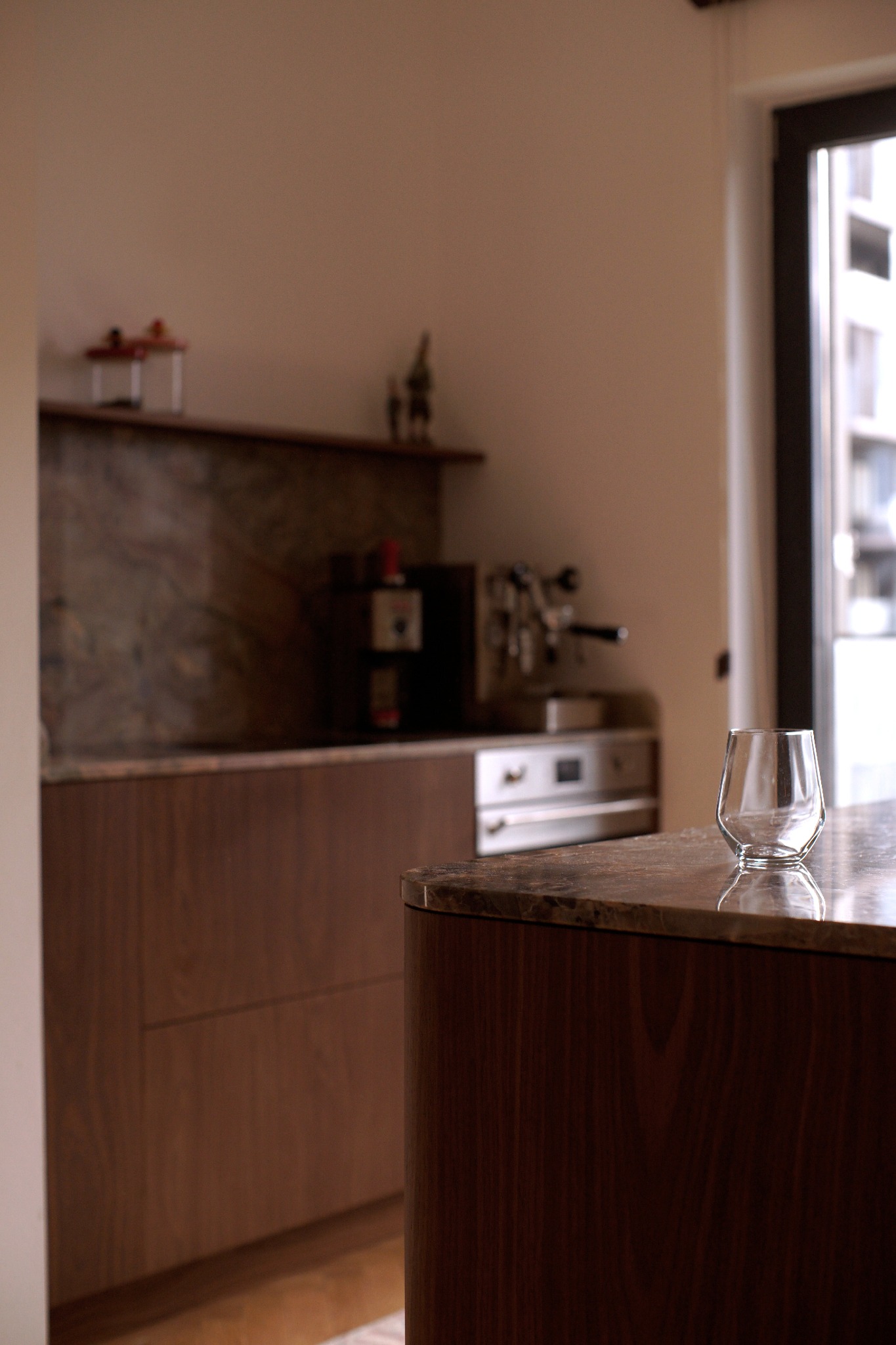 Nowoczesna kuchnia z drewnianymi frontami i marmurowym blatem. Na blacie stoi szklanka, w tle ekspres do kawy i okno z widokiem na budynek.