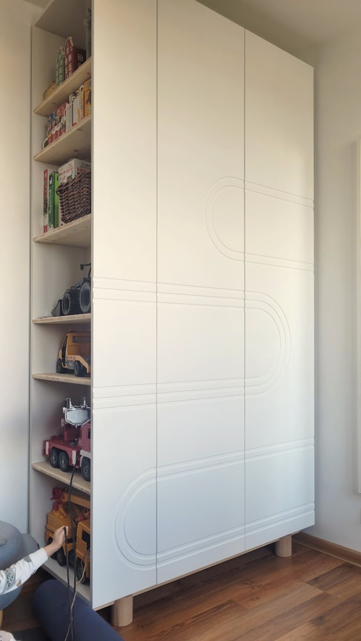 Biała szafa z półkami, wypełniona zabawkami, z dekoracyjnym frezem na frontach, stojąca na drewnianych nóżkach w pokoju dziecięcym.