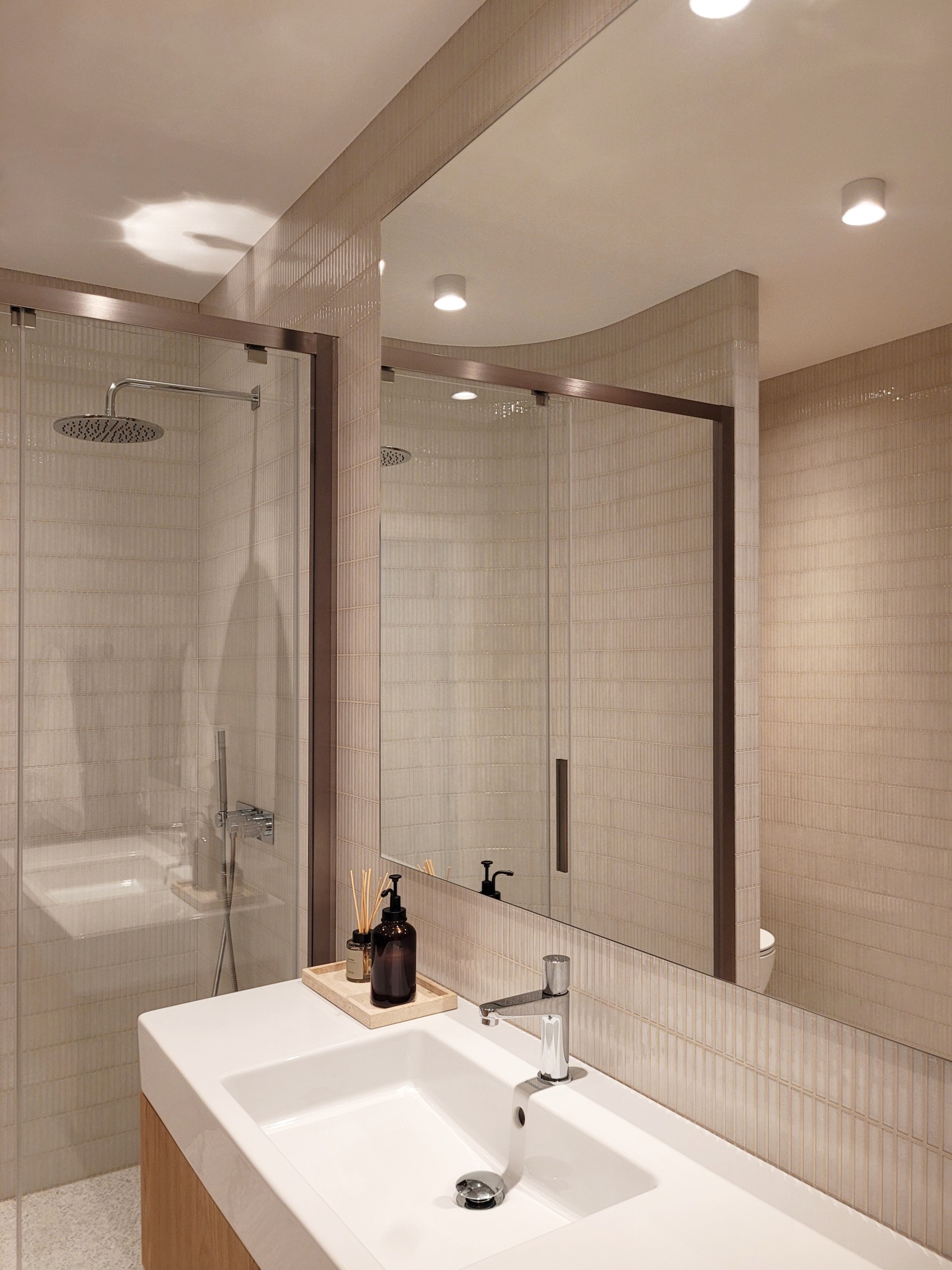 Nowoczesna łazienka z kabiną prysznicową typu walk-in, białą umywalką nablatową i dużym lustrem. Beżowe płytki na ścianach dodają ciepła wnętrzu.