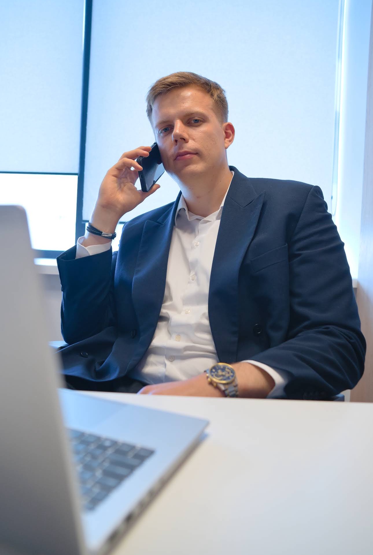 Doradca finansowy w marynarce rozmawia przez telefon w biurze, widoczny laptop i zegarek na ręce. Profesjonalna obsługa klienta w trakcie rozmowy.