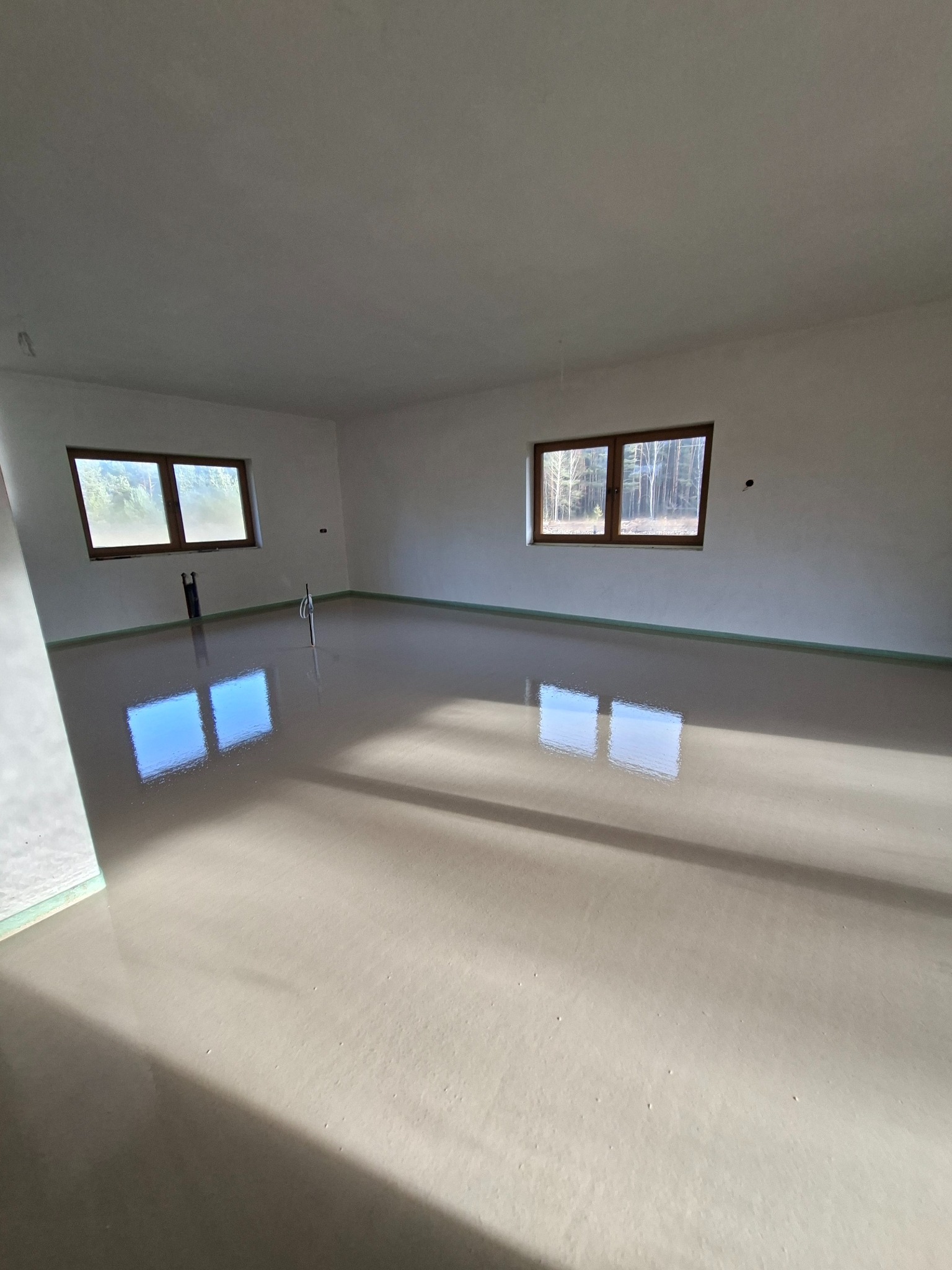 Jasne wnętrze z gładką, szarą posadzką, odbijającą światło z okien. Widoczne pionowe rury i listwy przypodłogowe. Minimalistyczny design, świeżo wylana powierzchnia.