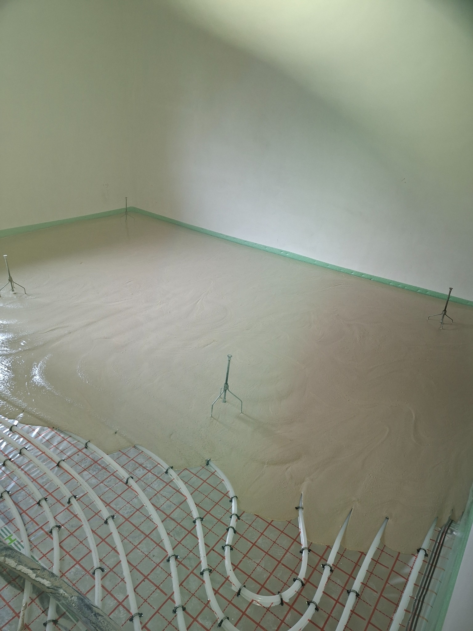 Jasna, świeżo wylana posadzka z widocznymi rurami ogrzewania podłogowego i listwami dylatacyjnymi przy ścianach. Widoczne metalowe statywy poziomujące.