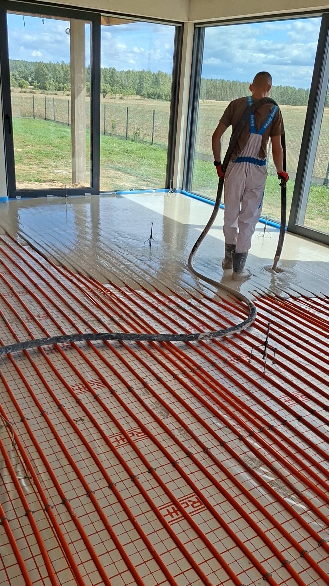 Wylewanie posadzki na ogrzewanie podłogowe w nowoczesnym wnętrzu z dużymi oknami. Widoczny pracownik w kombinezonie i czerwonych rękawicach podczas pracy.