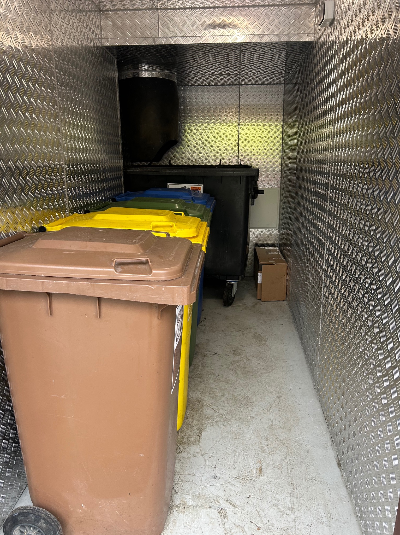 Wnętrze magazynu z rzędem kolorowych pojemników na odpady i kartonami, ściany wyłożone blachą ryflowaną. Widoczny duży, czarny kontener na kółkach.