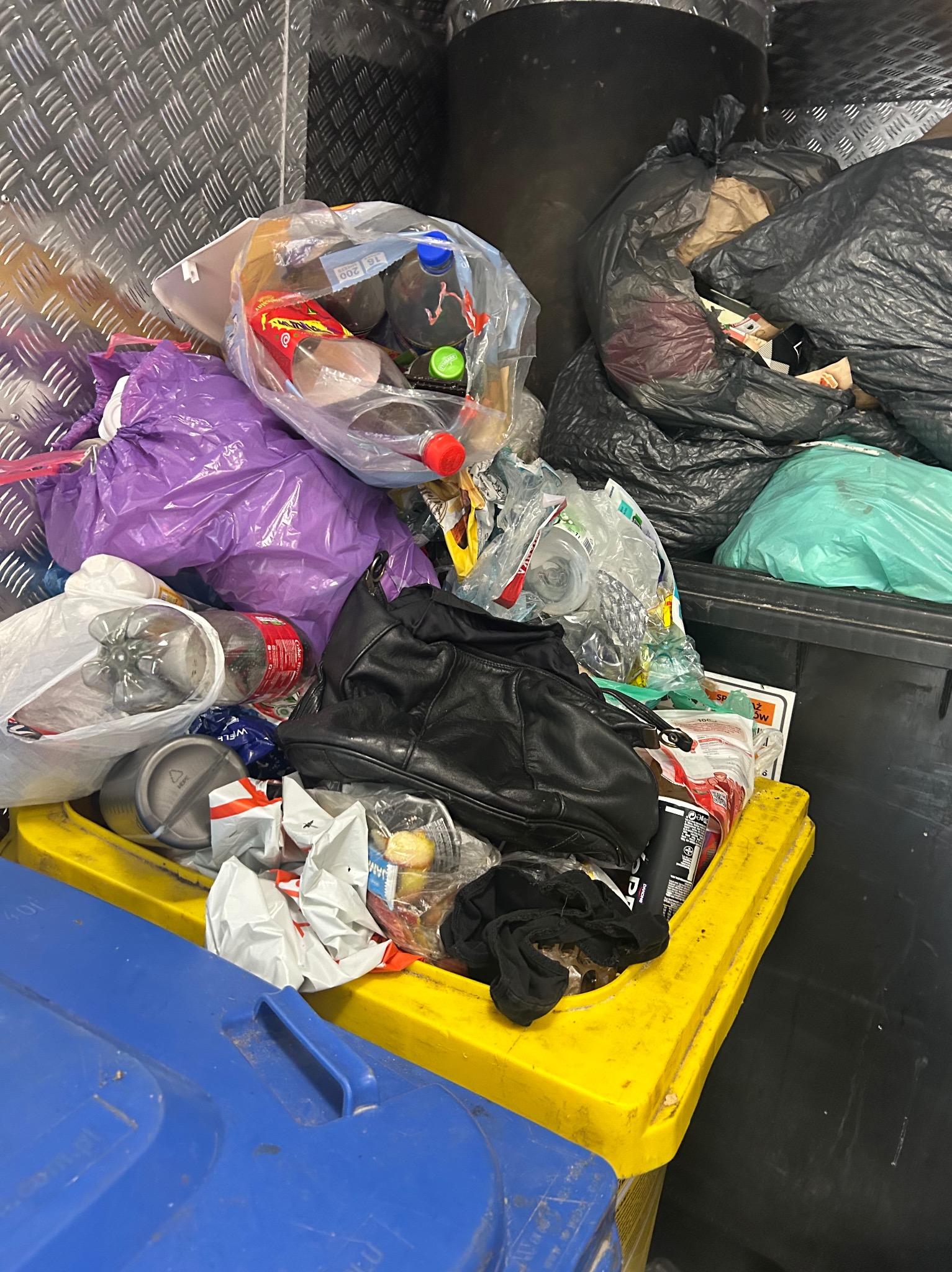 Pełen śmieci kontener z workami, butelkami i odpadami różnego rodzaju, ukazujący potrzebę uporządkowania i wyczyszczenia przestrzeni.