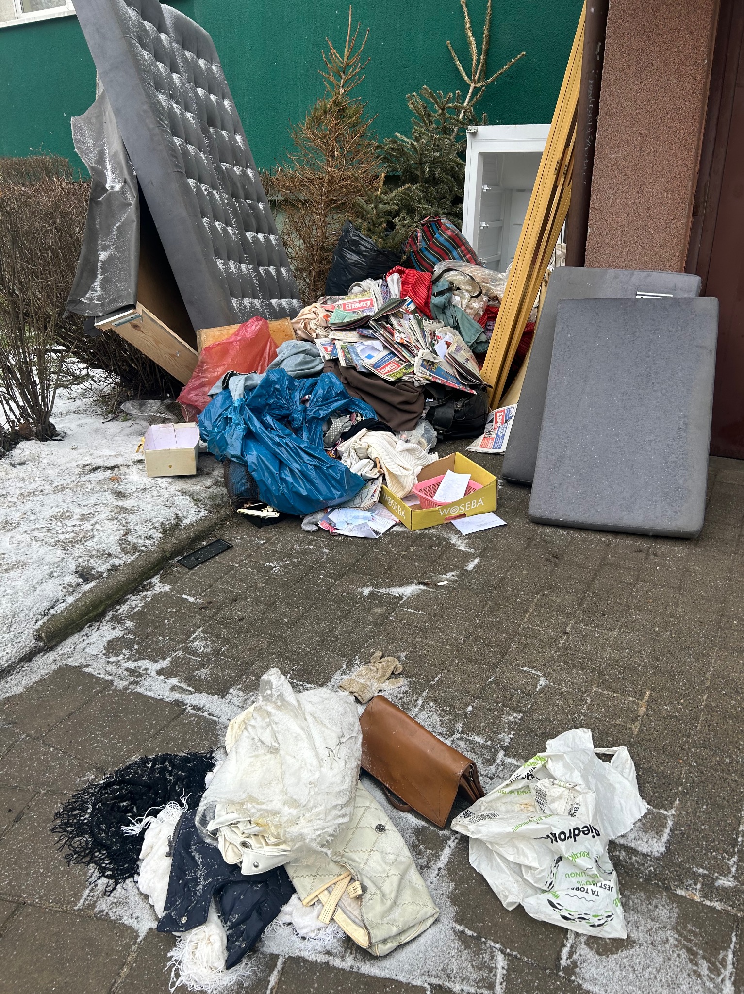 Sterta odpadów: materac, meble, ubrania, gazety i sprzęt AGD, porzucone na zewnątrz budynku w zimowej scenerii. Ślady śniegu na chodniku i przedmiotach.