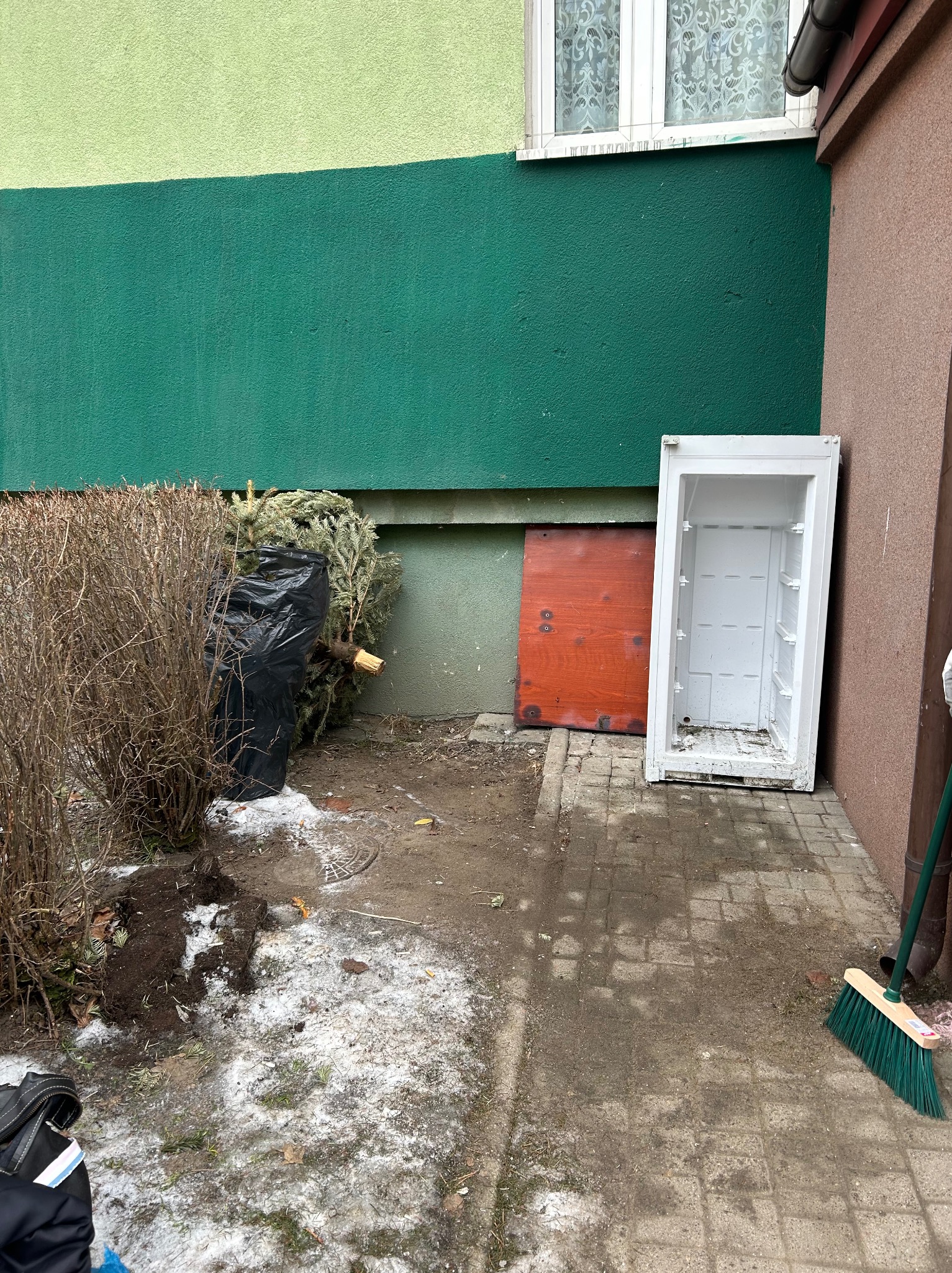Zewnętrzny teren po zimie: resztki śniegu, śmieci w worku, choinka, stara obudowa lodówki, deska i zielona miotła przy ścianie budynku.