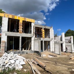 EMPRE Construction - Wznoszenie dwukondygnacyjnego domu z białego bloczka, widoczne szalunki okienne i stropowe. Na pierwszym planie gruz i deski, w tle błękitne niebo z chmurami.