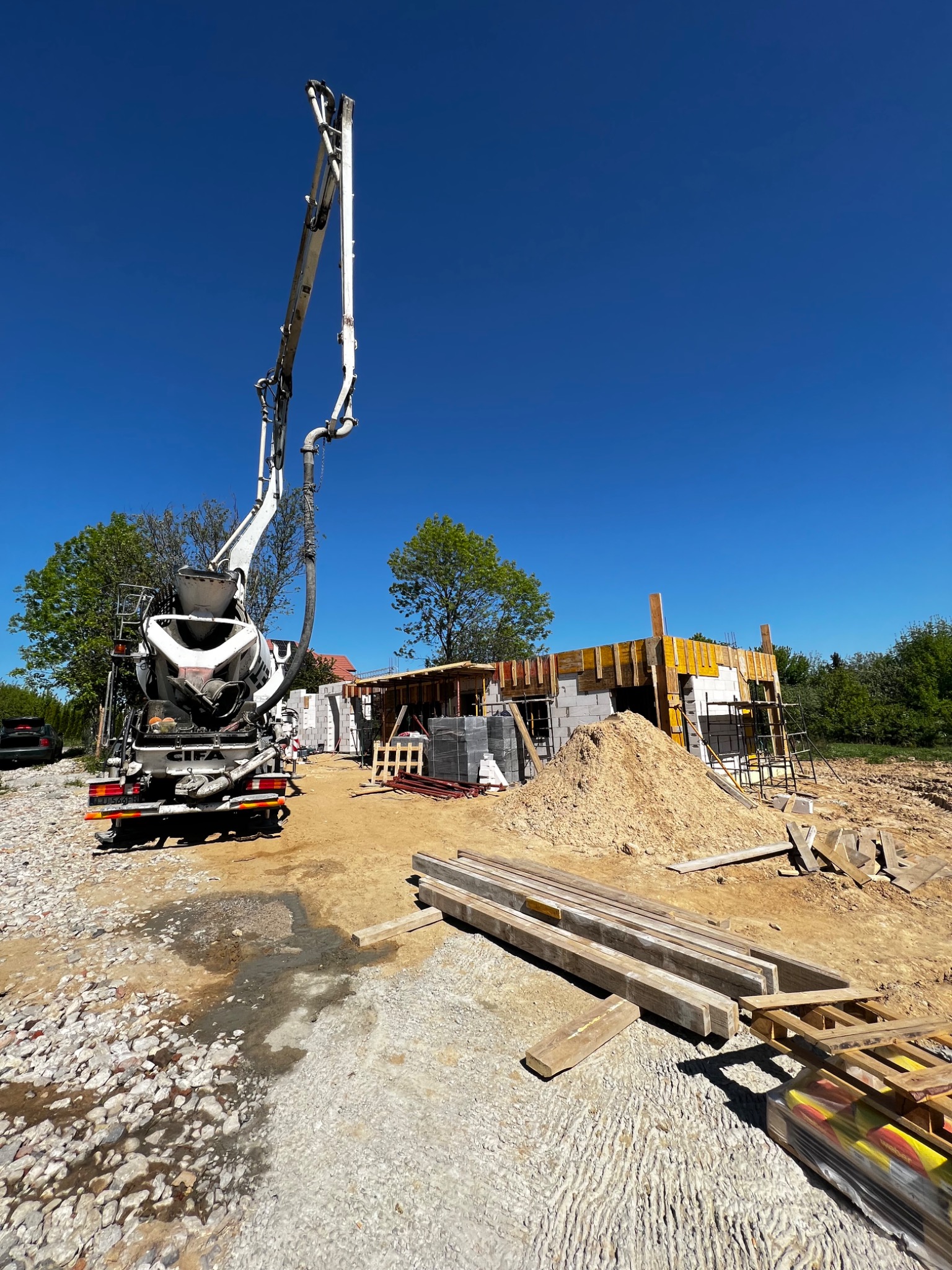 Betonomieszarka z pompą na budowie domu z bloczków, widoczne szalunki i materiały budowlane. Błękitne niebo w tle.