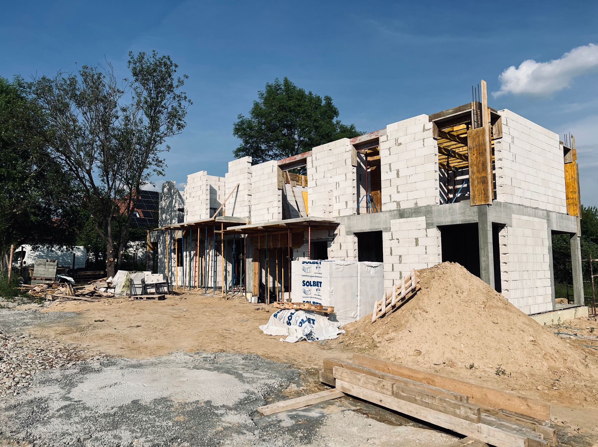 Wznoszenie ścian domu z białych bloczków Solbet, widoczne drewniane szalunki i zbrojenia. Na pierwszym planie hałda piasku i paleta z bloczkami. Słoneczny dzień.