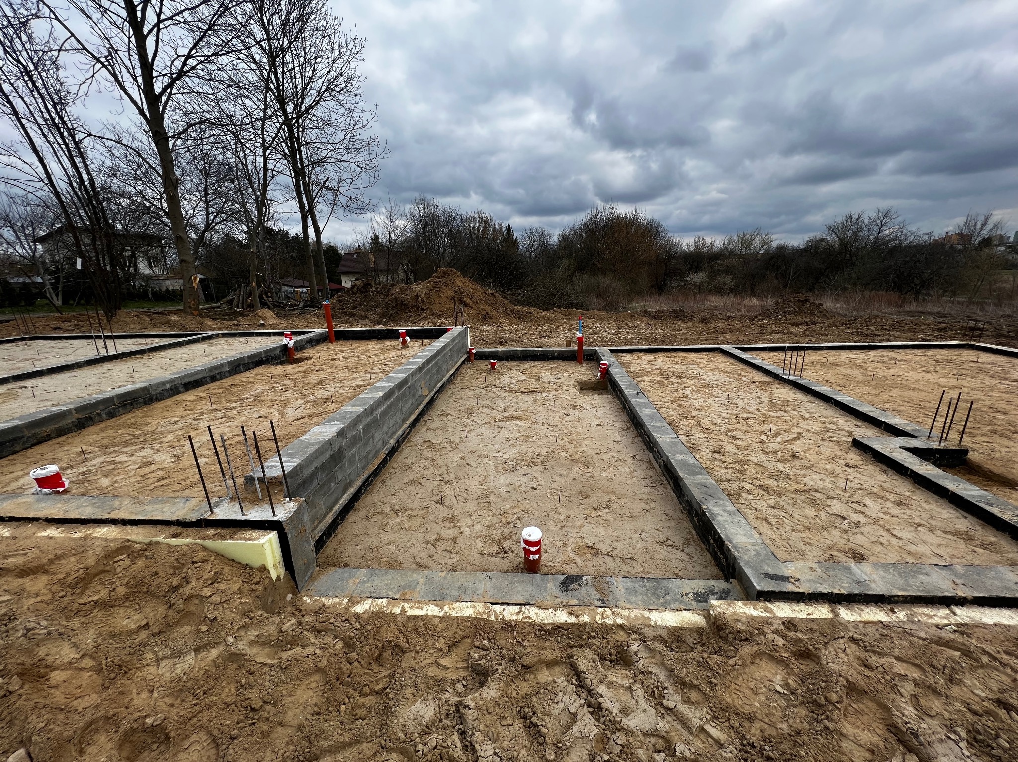 Zalane fundamenty pod budowę domu z widocznymi zbrojeniami i instalacją kanalizacyjną. Tło stanowią drzewa i pochmurne niebo.