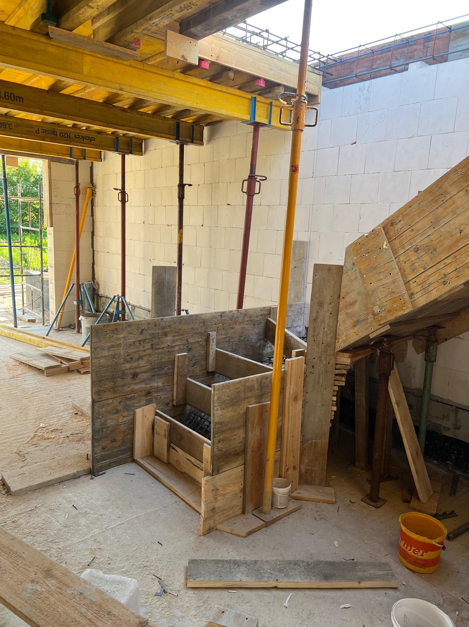 Szczegółowe ujęcie drewnianego szalunku schodów z widocznym zbrojeniem, wspartego na stemplach, w surowym wnętrzu budynku z bloczków.