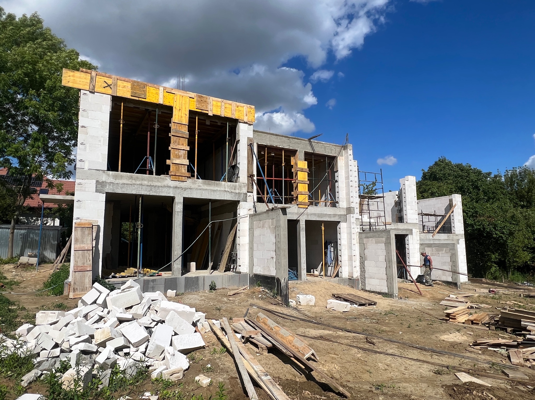 Wznoszenie dwukondygnacyjnego domu z białego bloczka, widoczne szalunki okienne i stropowe. Na pierwszym planie gruz i deski, w tle błękitne niebo z chmurami.