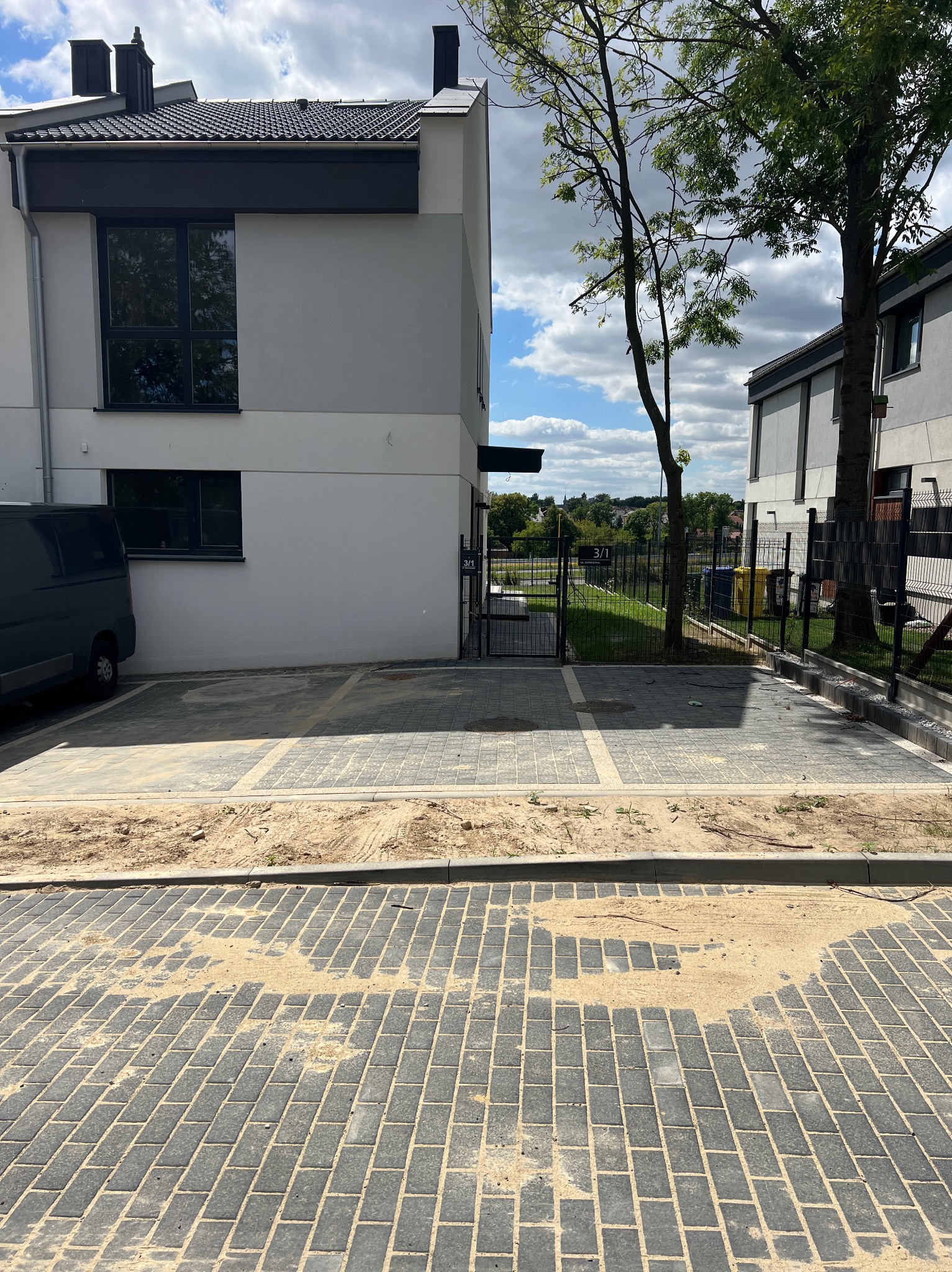 Nowoczesny dom bliźniak z szarą elewacją i brukowanym podjazdem, na którym widoczny piasek. Ujęcie z poziomu gruntu, w tle zieleń i błękitne niebo.