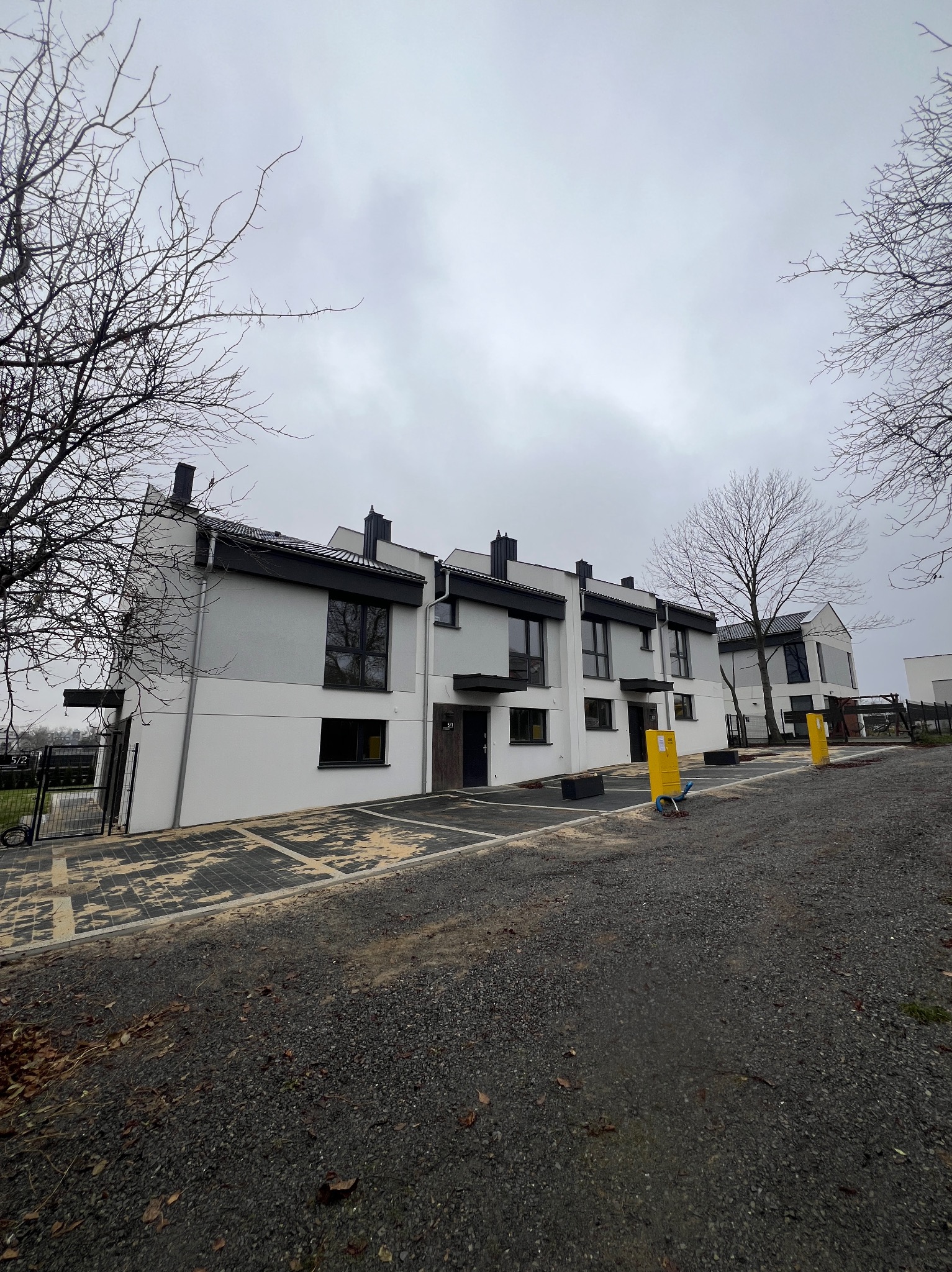 Nowoczesne, szeregowe domy z białymi elewacjami i grafitowymi akcentami, widoczne z perspektywy ulicy w pochmurny dzień. Żółte skrzynki gazowe na pierwszym planie.