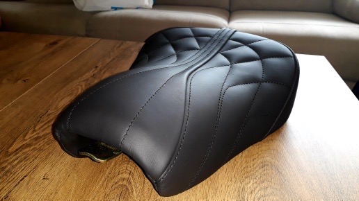 Czarne, pikowane siedzisko motocyklowe z nową tapicerką, leżące na drewnianym stole. Widoczne precyzyjne szwy i faktura skóry. W tle beżowa kanapa.