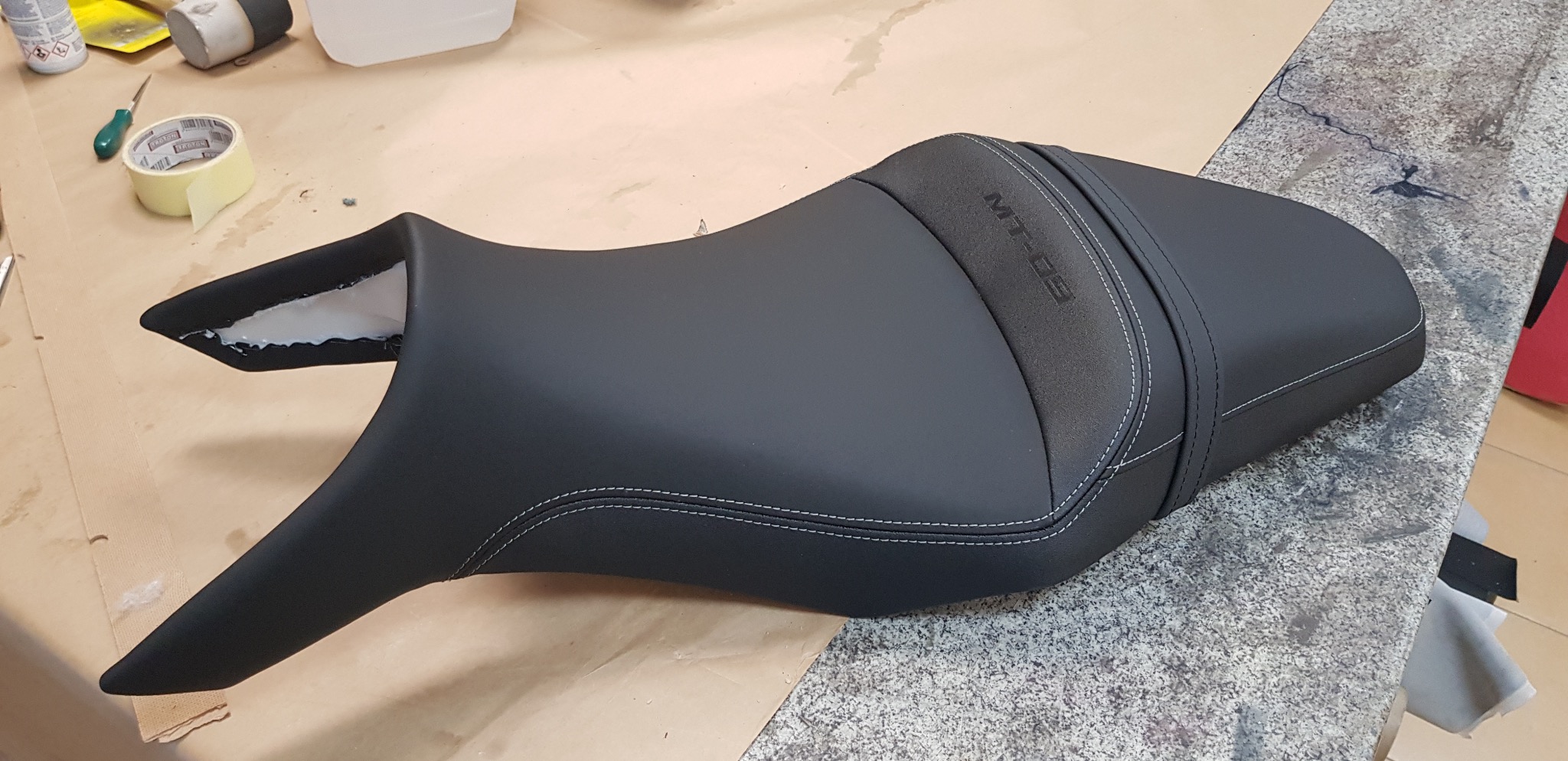 Czarne, odnowione siedzenie motocyklowe MT-09 z precyzyjnym, białym szwem, leżące na warsztatowym stole. Widoczne ślady kleju i narzędzia.