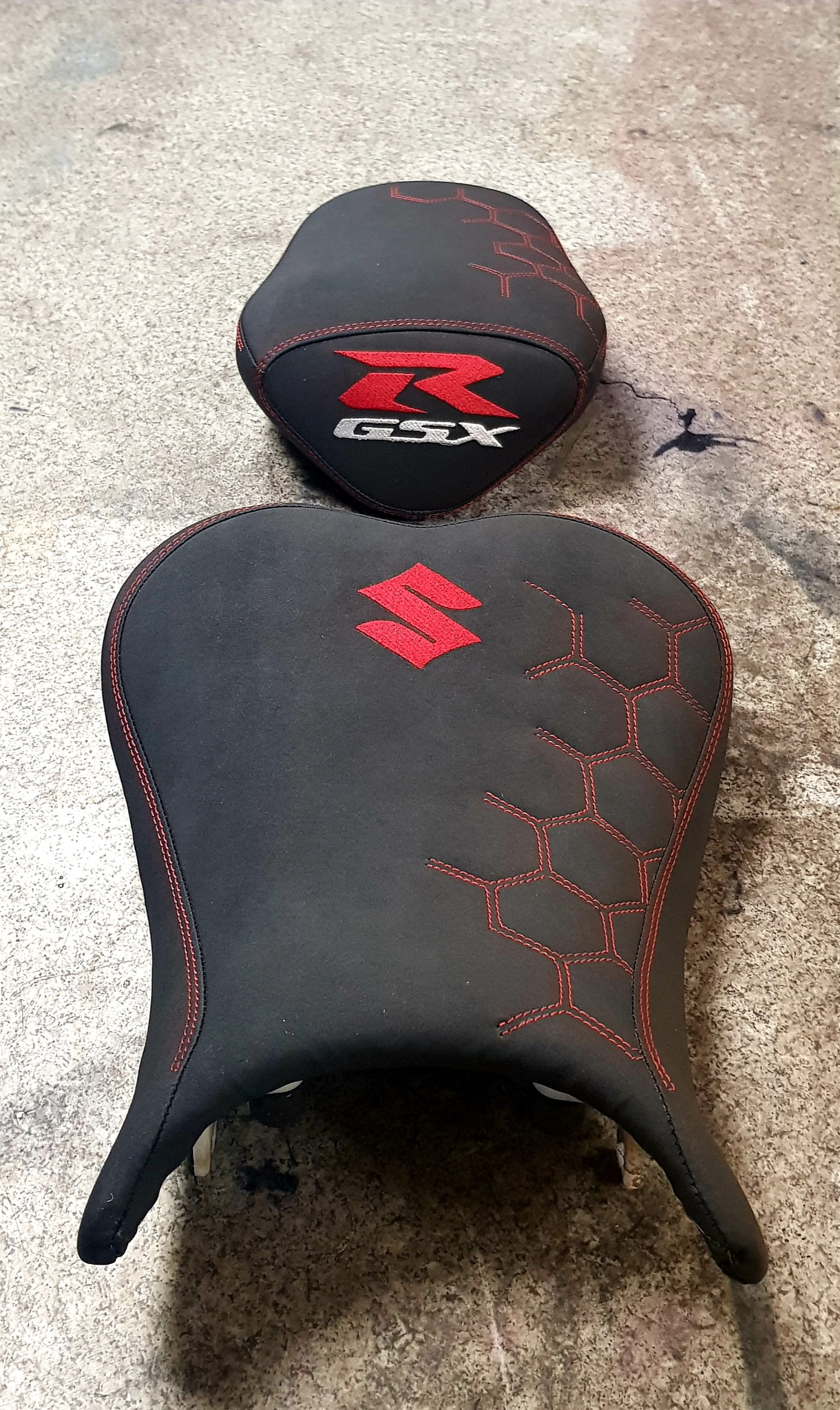 Dwa czarne, tapicerowane siedzenia motocyklowe z czerwonymi przeszyciami i logo Suzuki GSX, leżące na szarej powierzchni. Widoczne detale wykończenia i wzór plastra miodu.