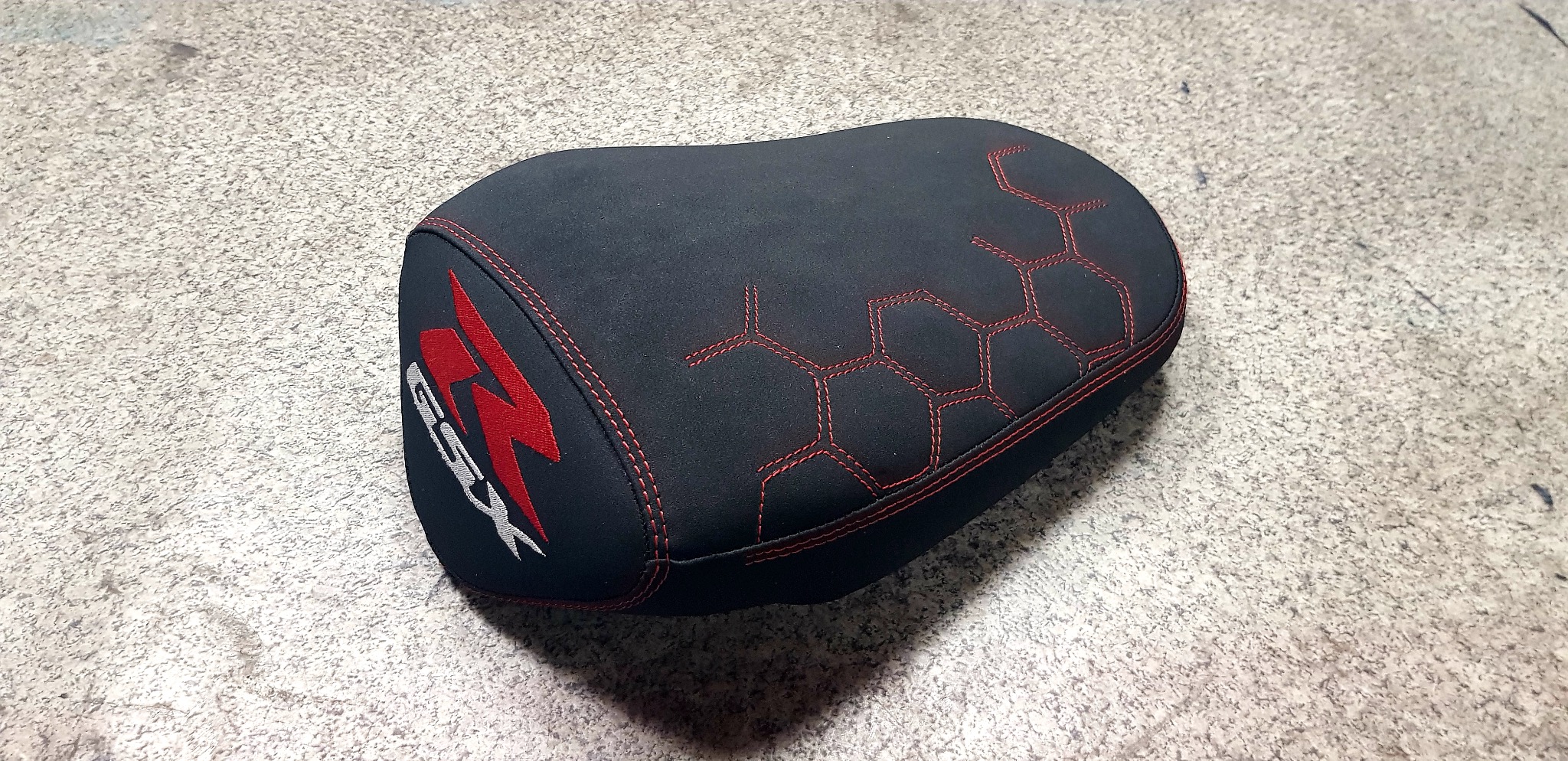 Czarne, odnowione siedzenie motocyklowe z czerwonymi przeszyciami i logo GSX, leżące na szarej powierzchni. Widoczne detale tapicerki i wzór sześciokątów.