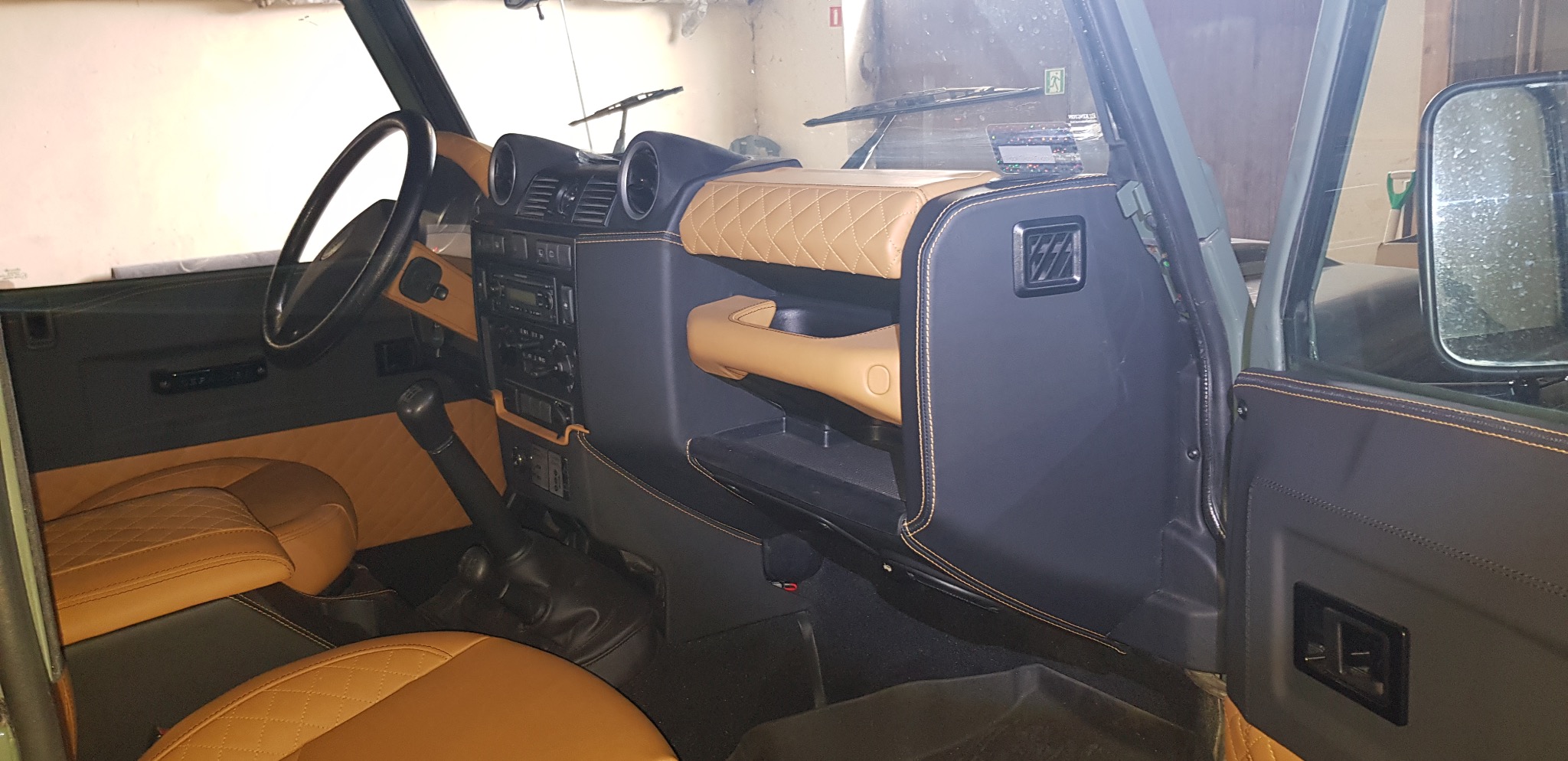 Wnętrze odrestaurowanego Land Rovera Defender z czarną tapicerką i beżowymi akcentami, w tym pikowane siedzenia i deska rozdzielcza, z widoczną kierownicą i dźwignią zmiany biegów.