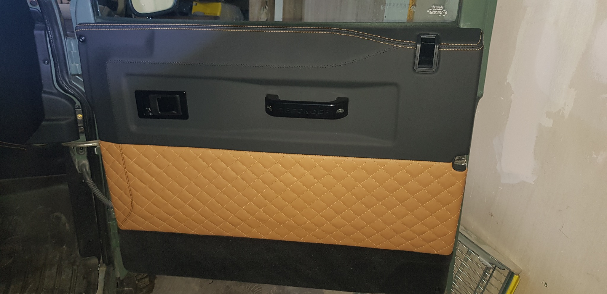 Odnowione wnętrze drzwi samochodu Defender z czarną górą i brązową, pikowaną skórą na dole, wykończone pomarańczową nicią. Detal tapicerski.