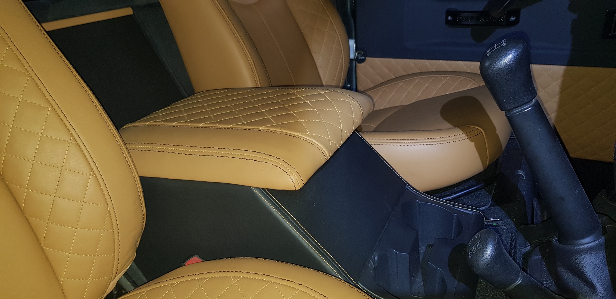Beżowa, pikowana tapicerka foteli samochodowych z czarnymi elementami wykończenia. Widoczna dźwignia zmiany biegów i hamulec ręczny. Detal wnętrza pojazdu.