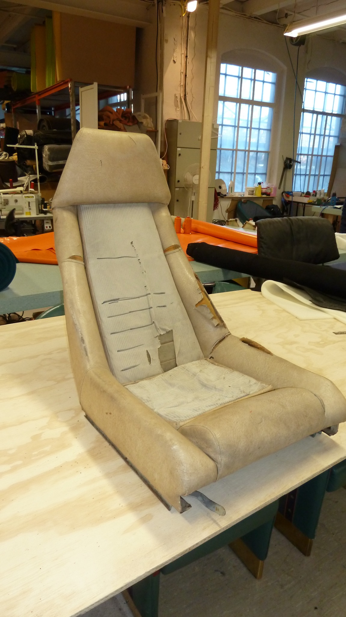 Zniszczone, beżowe siedzenie samochodowe z widocznymi przetarciami i dziurami na tapicerce, leżące na drewnianym stole w warsztacie tapicerskim.
