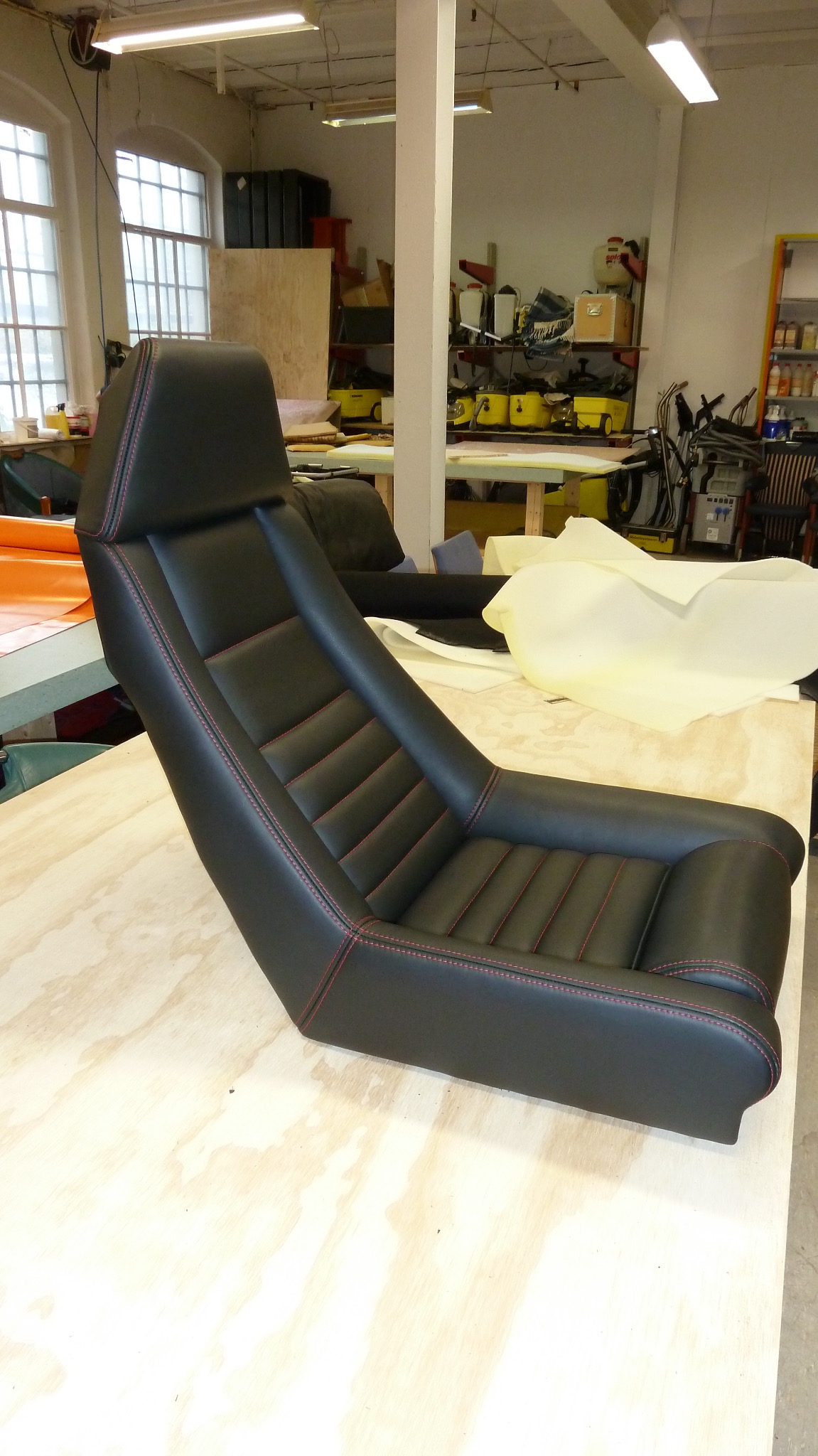 Odnowione czarne skórzane siedzenie samochodowe z czerwonymi przeszyciami, leżące na drewnianym stole w warsztacie tapicerskim. Widoczne materiały i narzędzia.